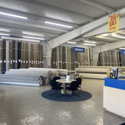 Carpet outlet showroom with neutral-toned rolls, a blue-topped service counter, consultation table, and a yellow 